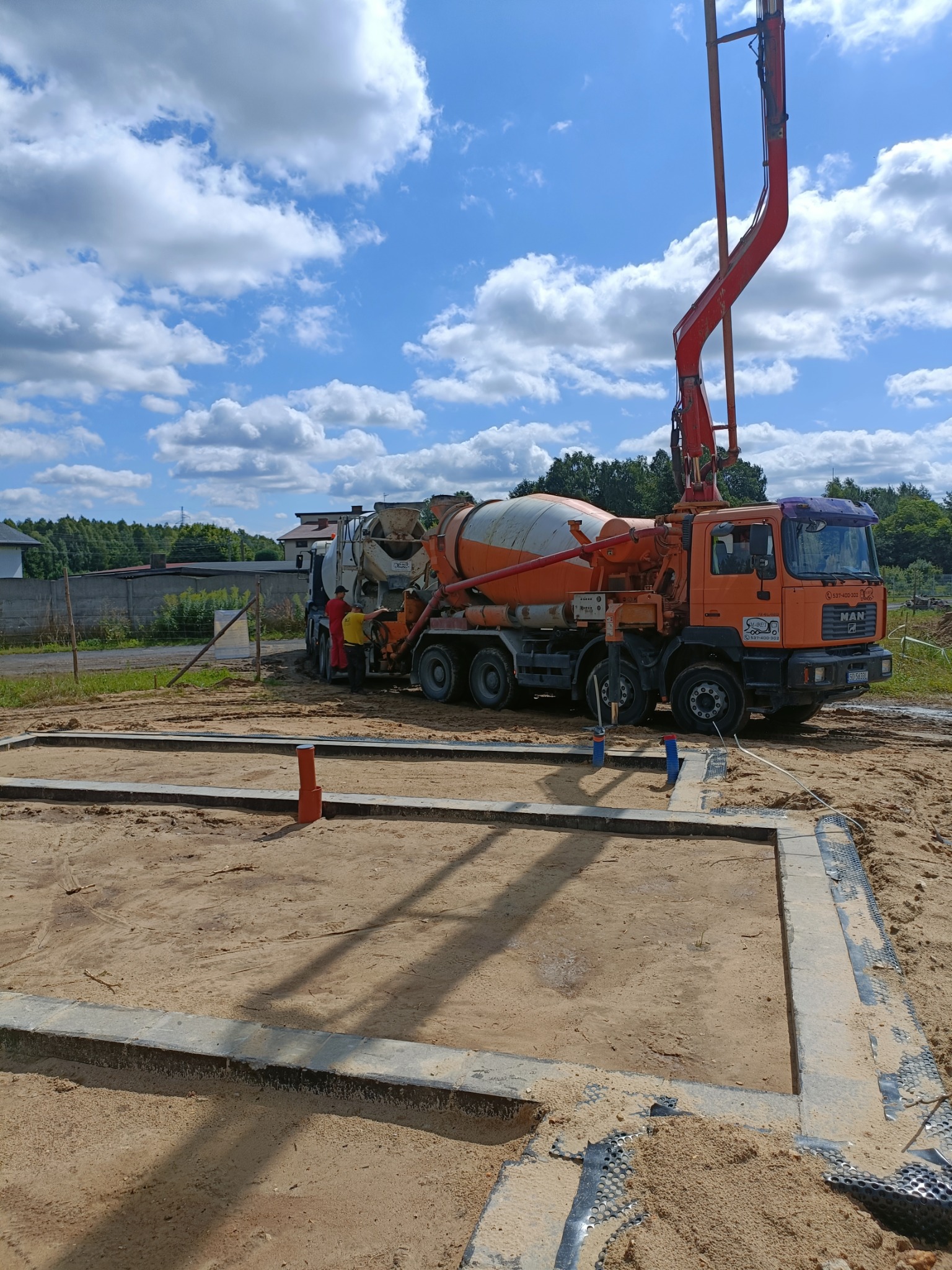Wylewanie betonu do szalunku fundamentów domu jednorodzinnego za pomocą pompy z betonomieszarki MAN. Na budowie dwóch pracowników w odzieży roboczej.