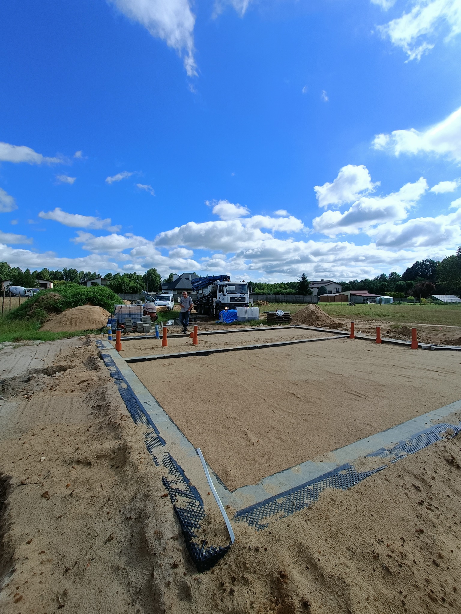 Przygotowany teren pod fundamenty domu jednorodzinnego. Widoczny obrys ław fundamentowych, piasek zasypowy, betonowy wieniec i pracownik nadzorujący prace.