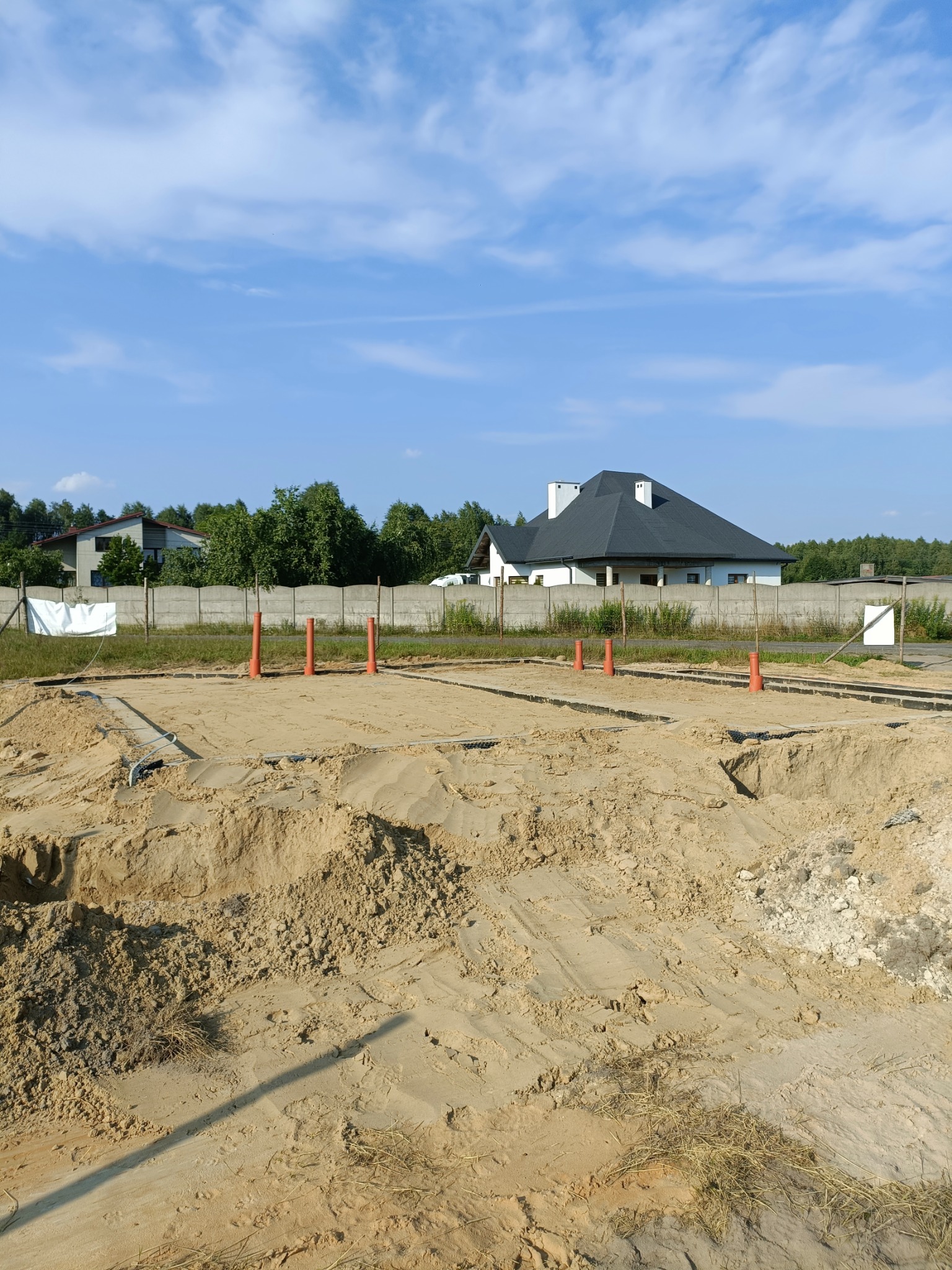 Przygotowany teren pod fundamenty domu jednorodzinnego z widocznymi wykopami, szalunkami i pionowymi rurami. W tle dom za betonowym ogrodzeniem.