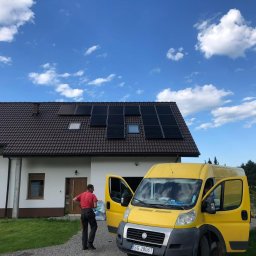 SMART PV ALEKSANDRA JABŁOŃSKA - Dom jednorodzinny z brązowym dachem, na którym zamontowano czarne panele słoneczne; na podjeździe stoi żółty bus z otwartymi drzwiami, obok którego stoi mężczyzna w czerwonej koszulce.