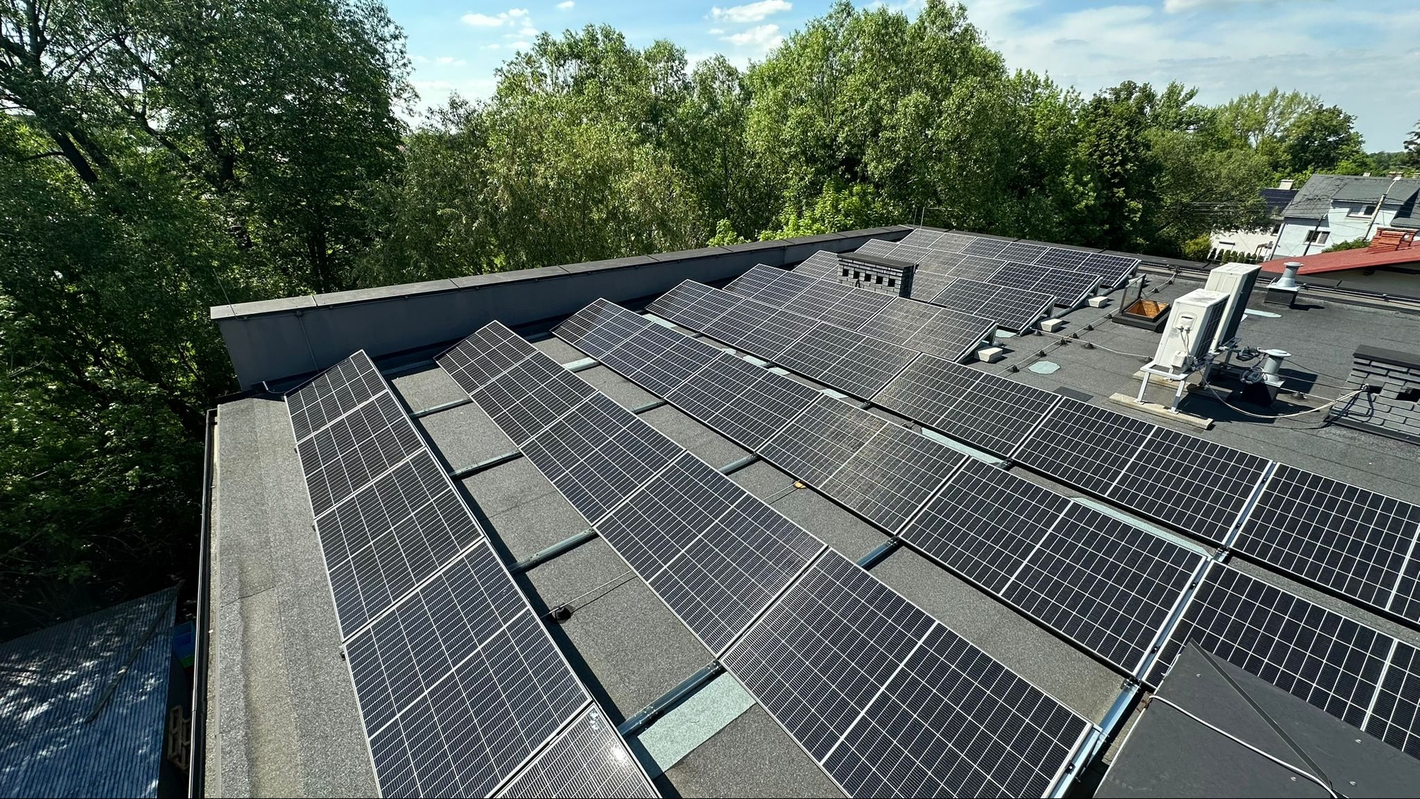 Widok z góry na instalację paneli słonecznych na płaskim dachu budynku, z widocznymi klimatyzatorami i zielenią drzew w tle pod błękitnym niebem.