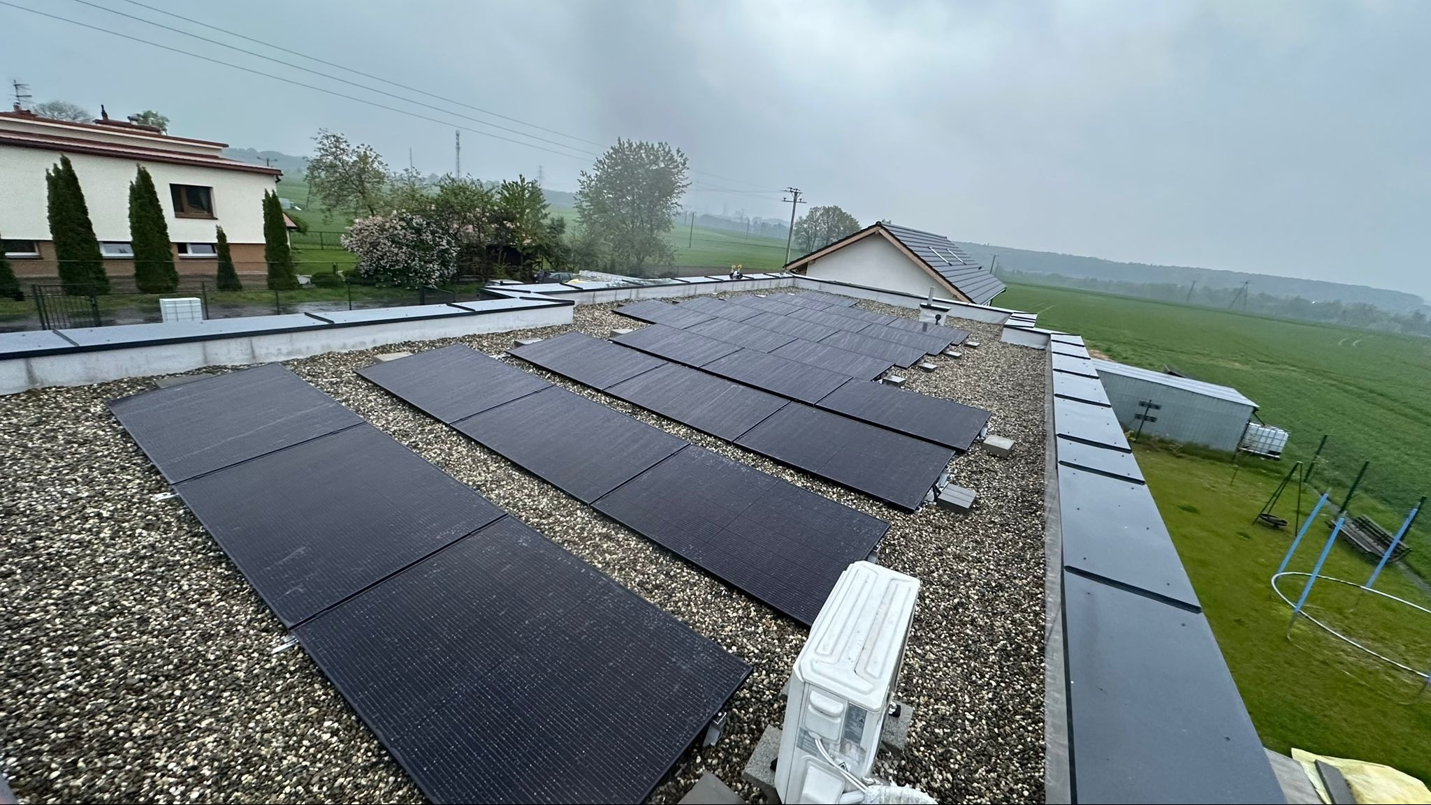 Widok z góry na instalację paneli słonecznych na płaskim dachu pokrytym żwirem, z widocznym budynkiem mieszkalnym i zielonymi polami w tle, uchwycony w pochmurny dzień.