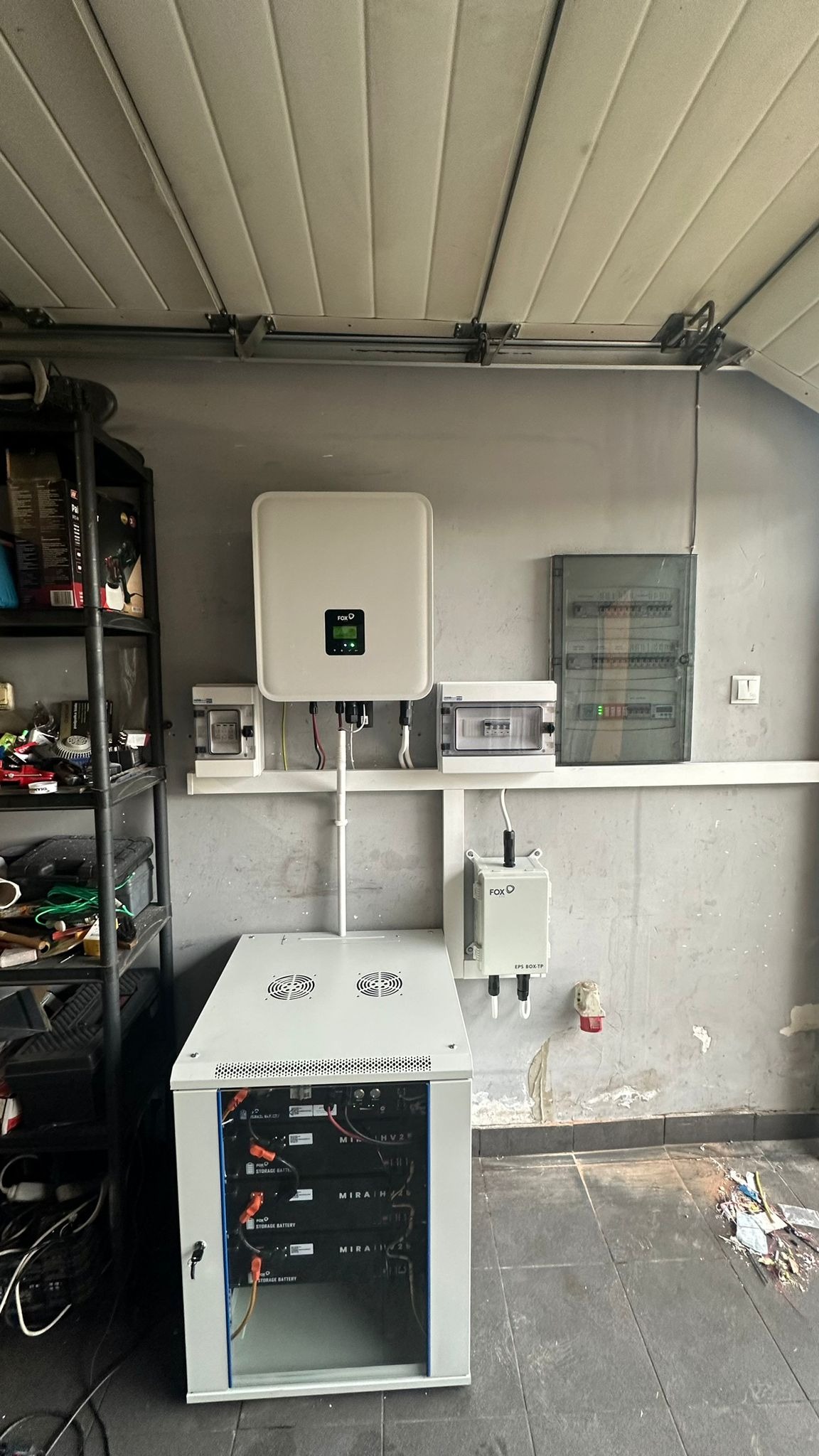 Kompletna instalacja fotowoltaiczna w garażu: inwerter FOX, moduł z bateriami Mira HV2, rozdzielnica elektryczna i okablowanie na szarej ścianie z widocznymi śladami użytkowania.