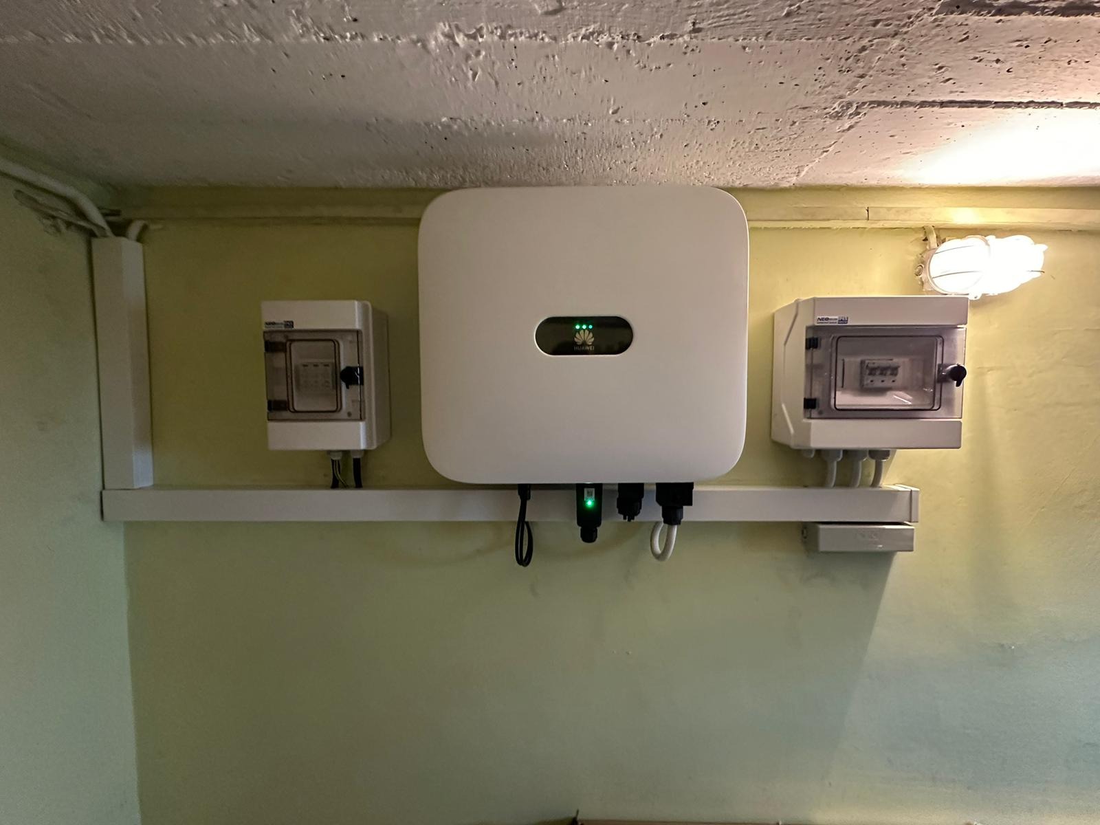 Biały inwerter fotowoltaiczny Huawei zamontowany na ścianie w pomieszczeniu, z widocznymi skrzynkami elektrycznymi po bokach i przewodami pod spodem, oświetlony lampą halogenową.