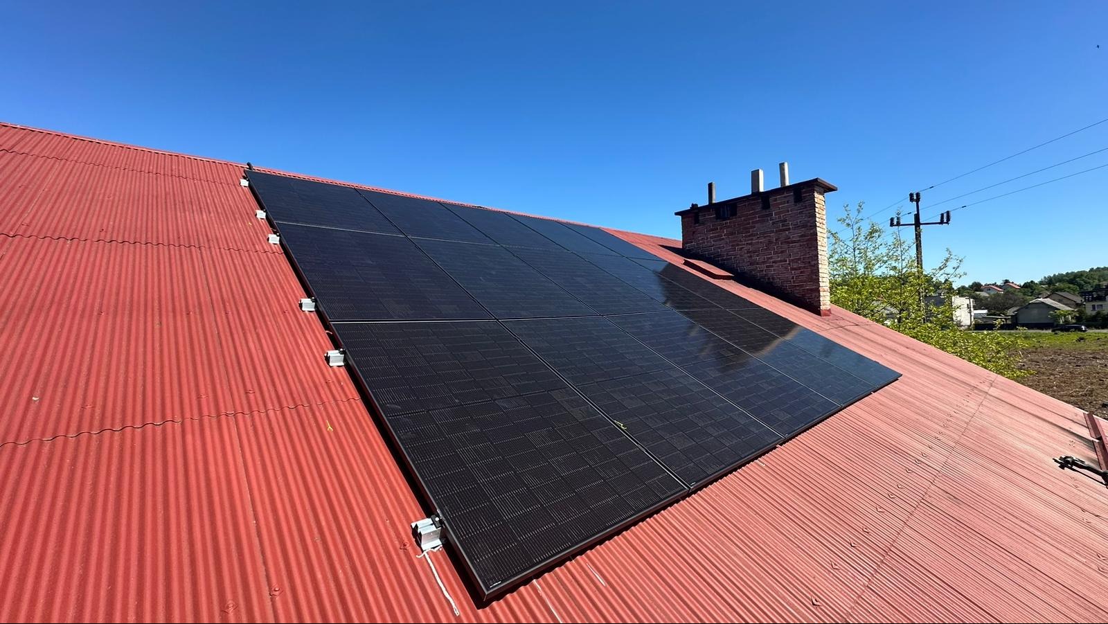 Panele słoneczne zamontowane na czerwonym, falistym dachu, z widocznym ceglanym kominem i błękitnym niebem w tle.