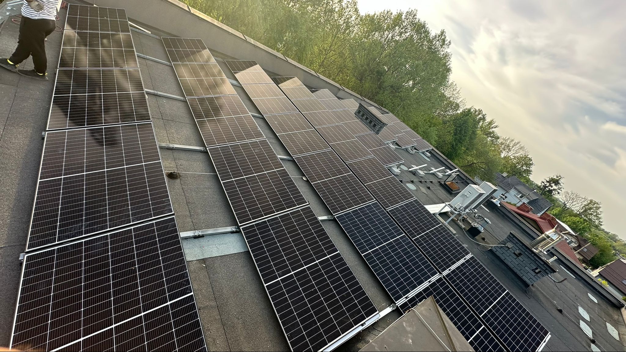 Instalacja fotowoltaiczna na płaskim dachu budynku mieszkalnego, widoczne panele słoneczne, elementy montażowe oraz fragment sylwetki osoby podczas montażu.