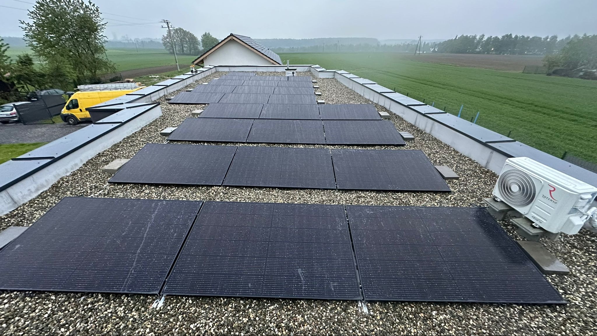 Panel słoneczny na dachu pokrytym żwirem, widoczny klimatyzator Rotenso i żółty samochód dostawczy w tle.