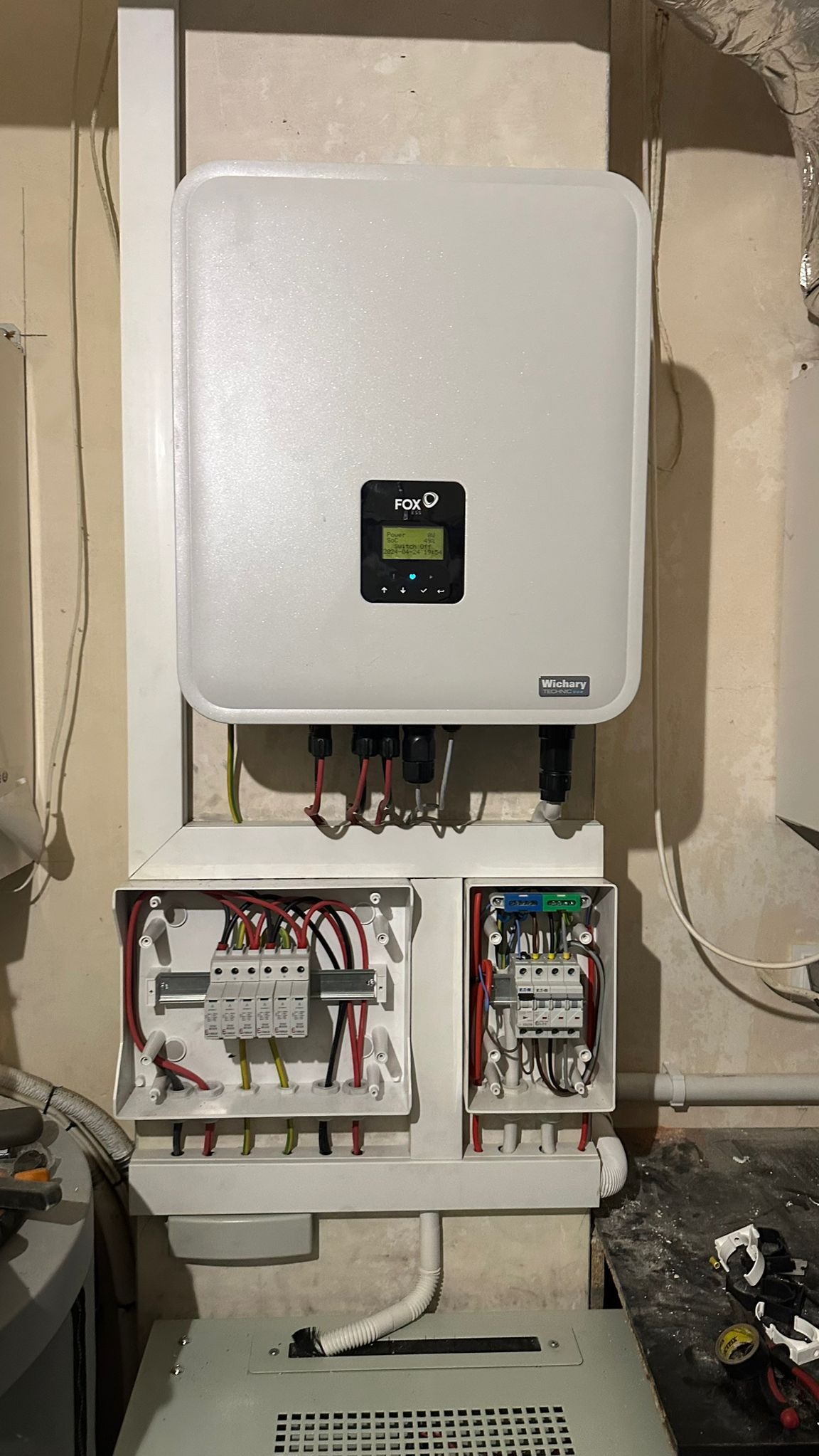 Biały inwerter fotowoltaiczny marki Fox zamontowany na ścianie, z widocznymi skrzynkami rozdzielczymi i okablowaniem, na tle surowej ściany.