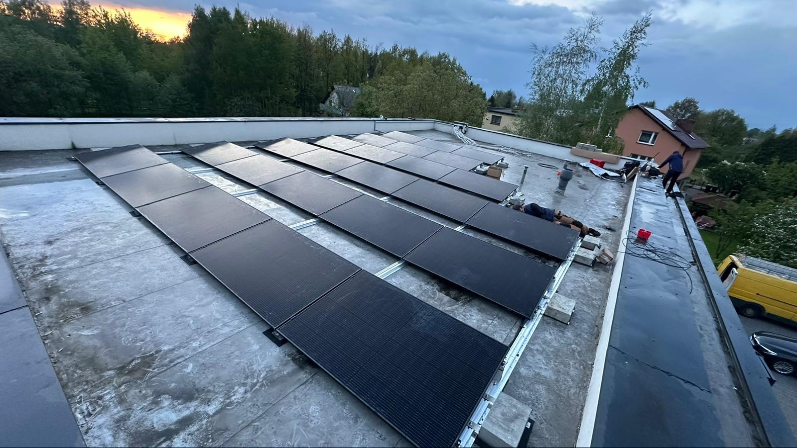 Montaż paneli fotowoltaicznych na płaskim dachu, widoczni dwaj pracownicy w trakcie instalacji, żółty samochód dostawczy zaparkowany w tle.