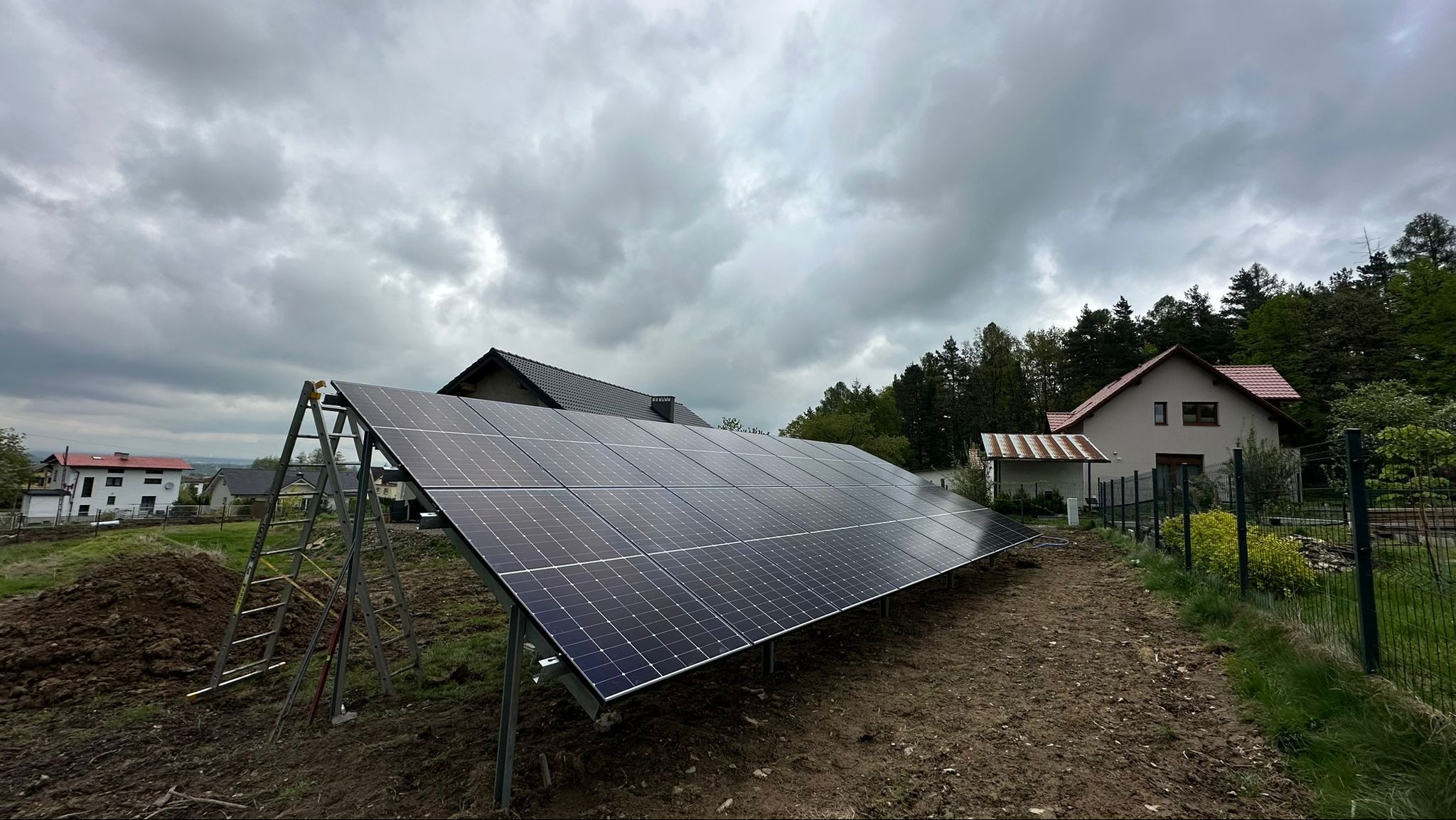 Panele słoneczne ustawione na metalowej konstrukcji na ziemi, drabina oparta o konstrukcję z lewej strony, w tle domy i pochmurne niebo.