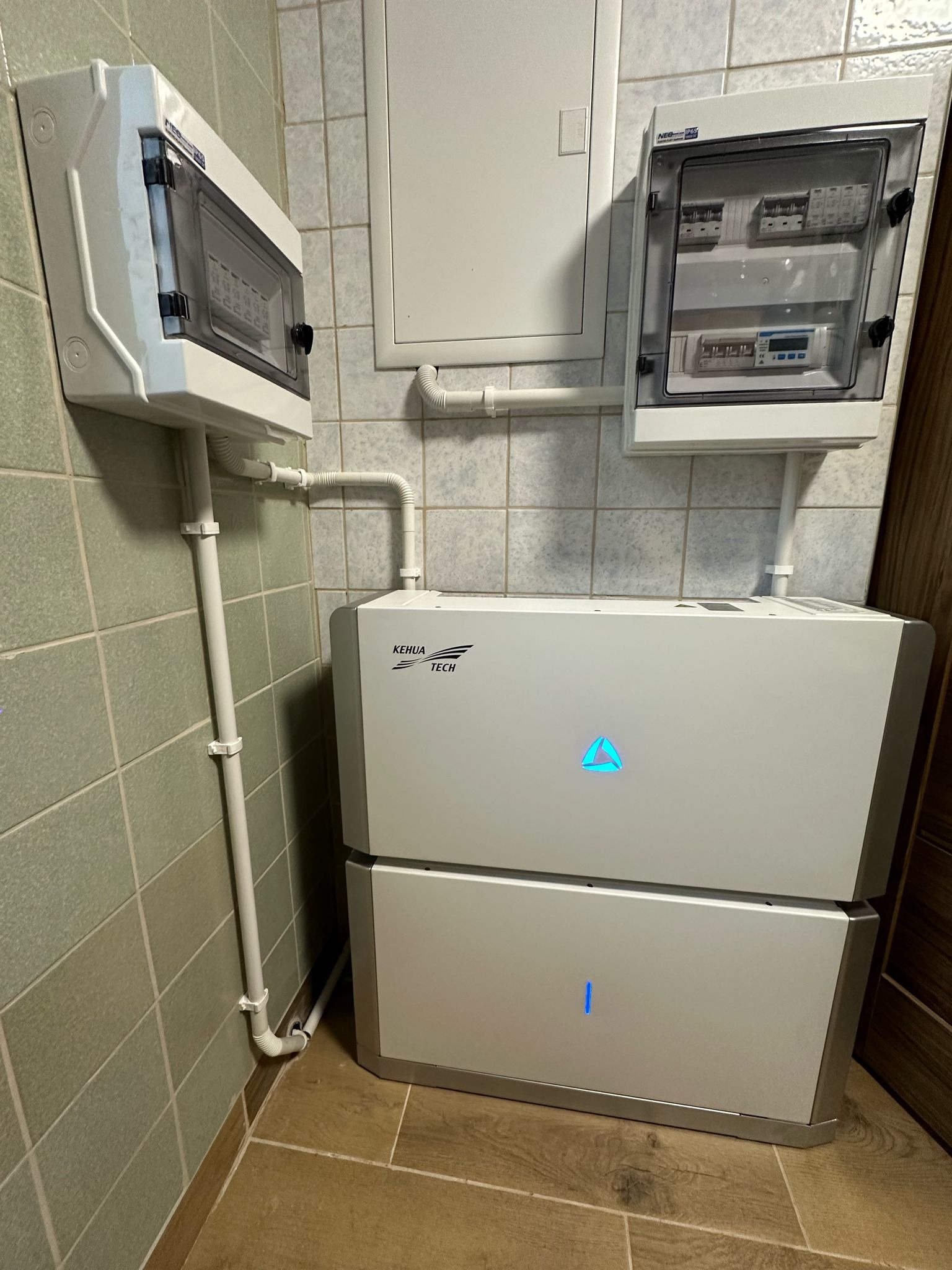 Biały, dwuczęściowy akumulator energii z logo Kehua Tech, zamontowany w pomieszczeniu z kafelkami, obok widoczne dwie skrzynki elektryczne i rury PCV.