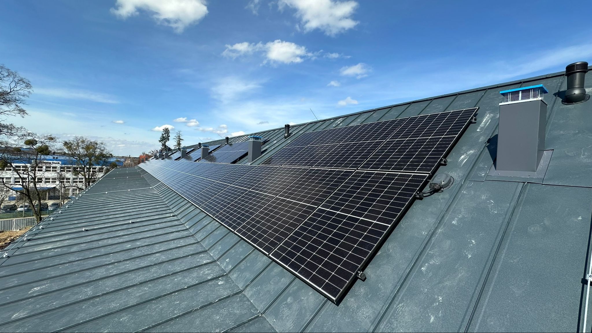 Panele słoneczne na metalowym dachu z kominami wentylacyjnymi, błękitne niebo w tle.