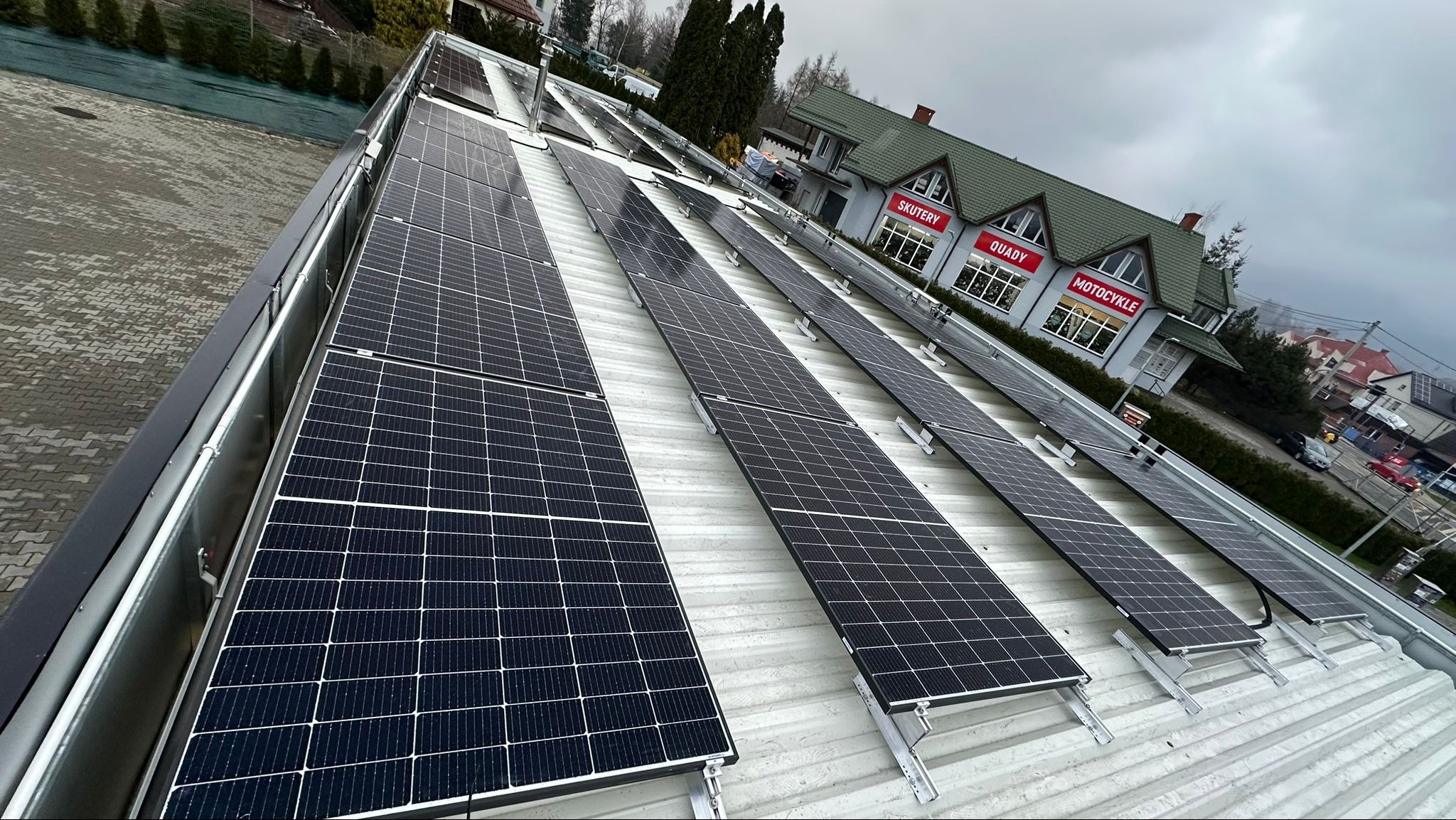 Rząd paneli fotowoltaicznych zamontowanych na metalowym dachu budynku komercyjnego, widok z góry pod kątem, w tle budynek z szyldem 'Skutery, Quady, Motocykle'.