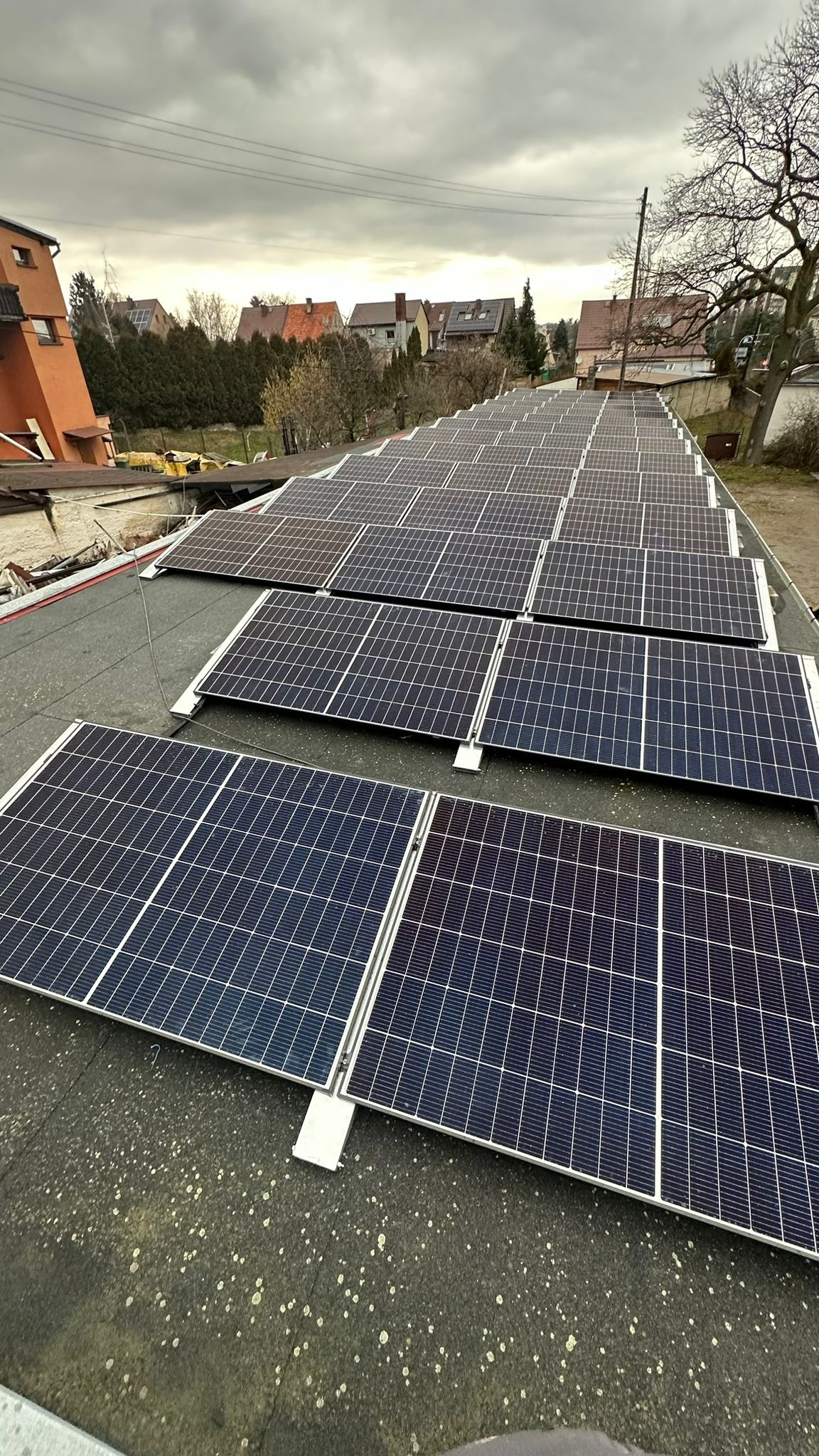 Widok z góry na kompletną instalację paneli słonecznych na płaskim dachu budynku mieszkalnego, z widocznymi sąsiednimi domami i pochmurnym niebem w tle.