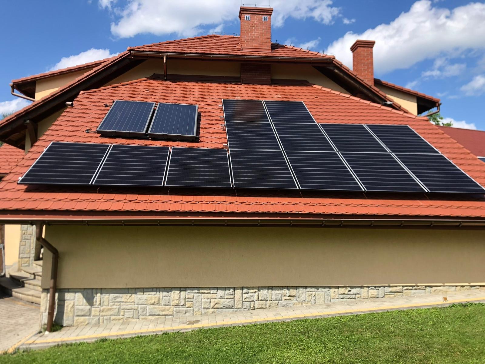 Panele słoneczne w dwóch sekcjach na dachu domu z czerwonej dachówki, widoczne kominy i błękitne niebo.