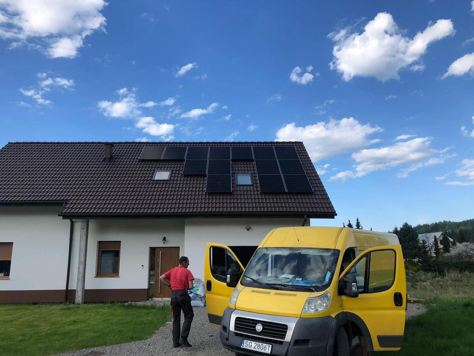 Dom jednorodzinny z brązowym dachem, na którym zamontowano czarne panele słoneczne; na podjeździe stoi żółty bus z otwartymi drzwiami, obok którego stoi mężczyzna w czerwonej koszulce.