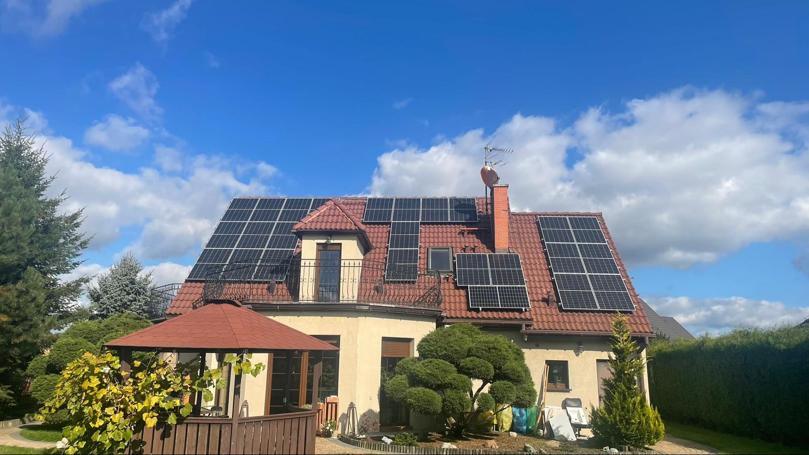 Dom jednorodzinny z czerwoną dachówką i panelami fotowoltaicznymi zamontowanymi na dachu, widok z poziomu ogrodu w słoneczny dzień.