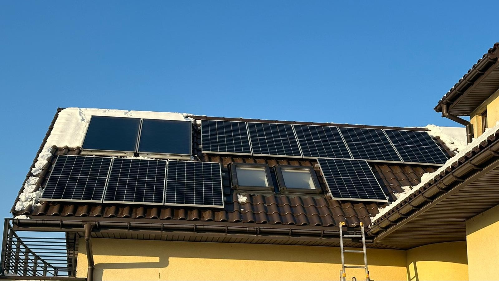 Panele fotowoltaiczne i kolektory słoneczne na brązowym dachu pokrytym częściowo śniegiem, drabina oparta o ścianę budynku.