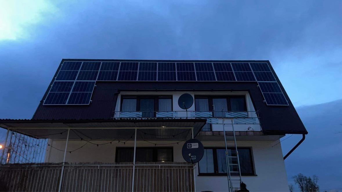 Instalacja fotowoltaiczna na dwuspadowym dachu domu jednorodzinnego, widoczne panele słoneczne w układzie prostokątnym, antena satelitarna na balkonie, drabina oparta o ścianę budynku, pochmurne...