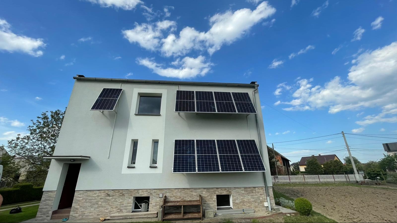 Panele słoneczne zamontowane na elewacji dwupiętrowego domu z jasną elewacją i kamienną podmurówką, na tle błękitnego nieba z białymi obłokami.