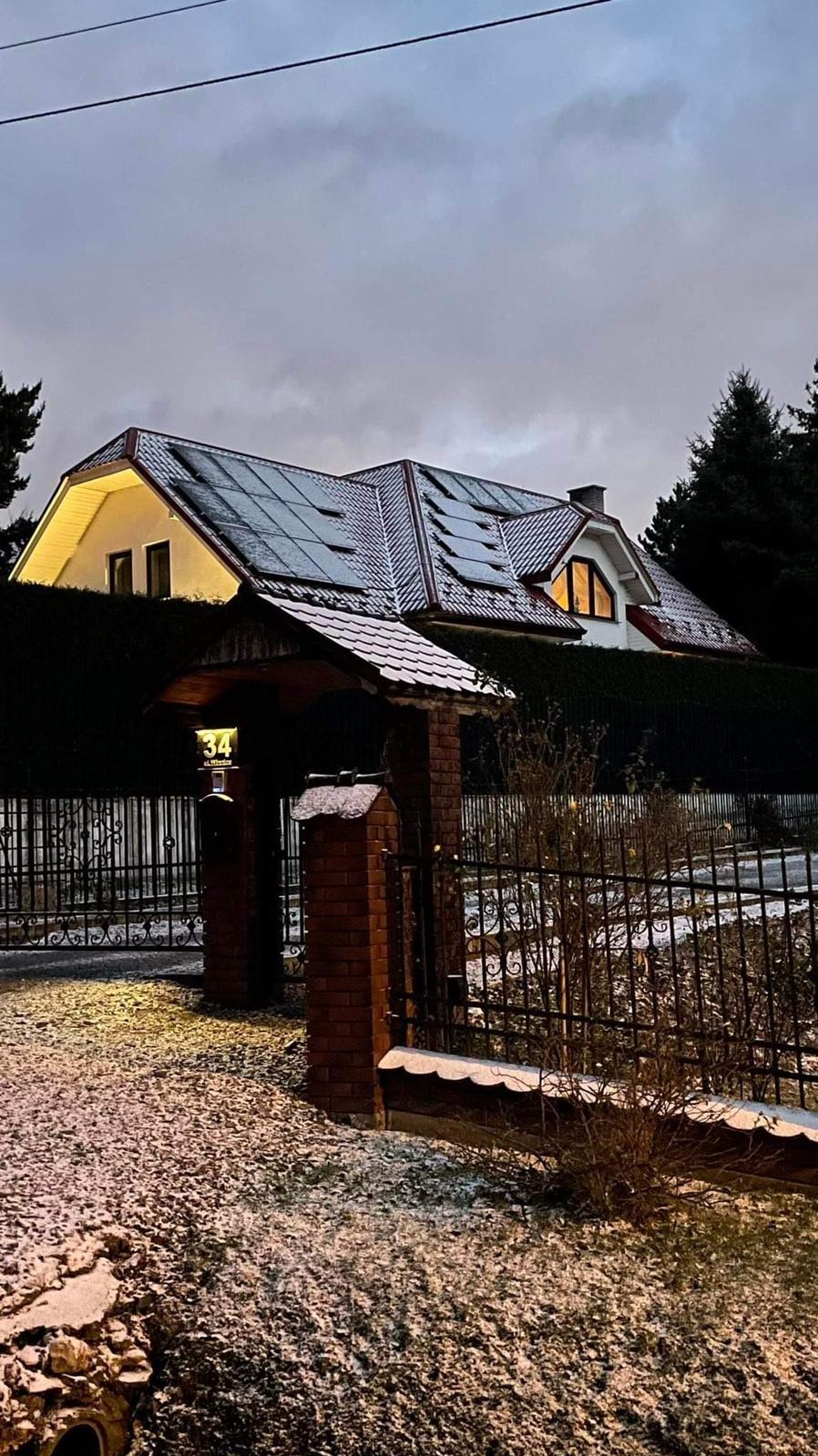Dom jednorodzinny z panelami fotowoltaicznymi na dachu, lekko przyprószonym śniegiem, widoczny od strony ulicy przez ozdobną bramę z numerem domu '34'.