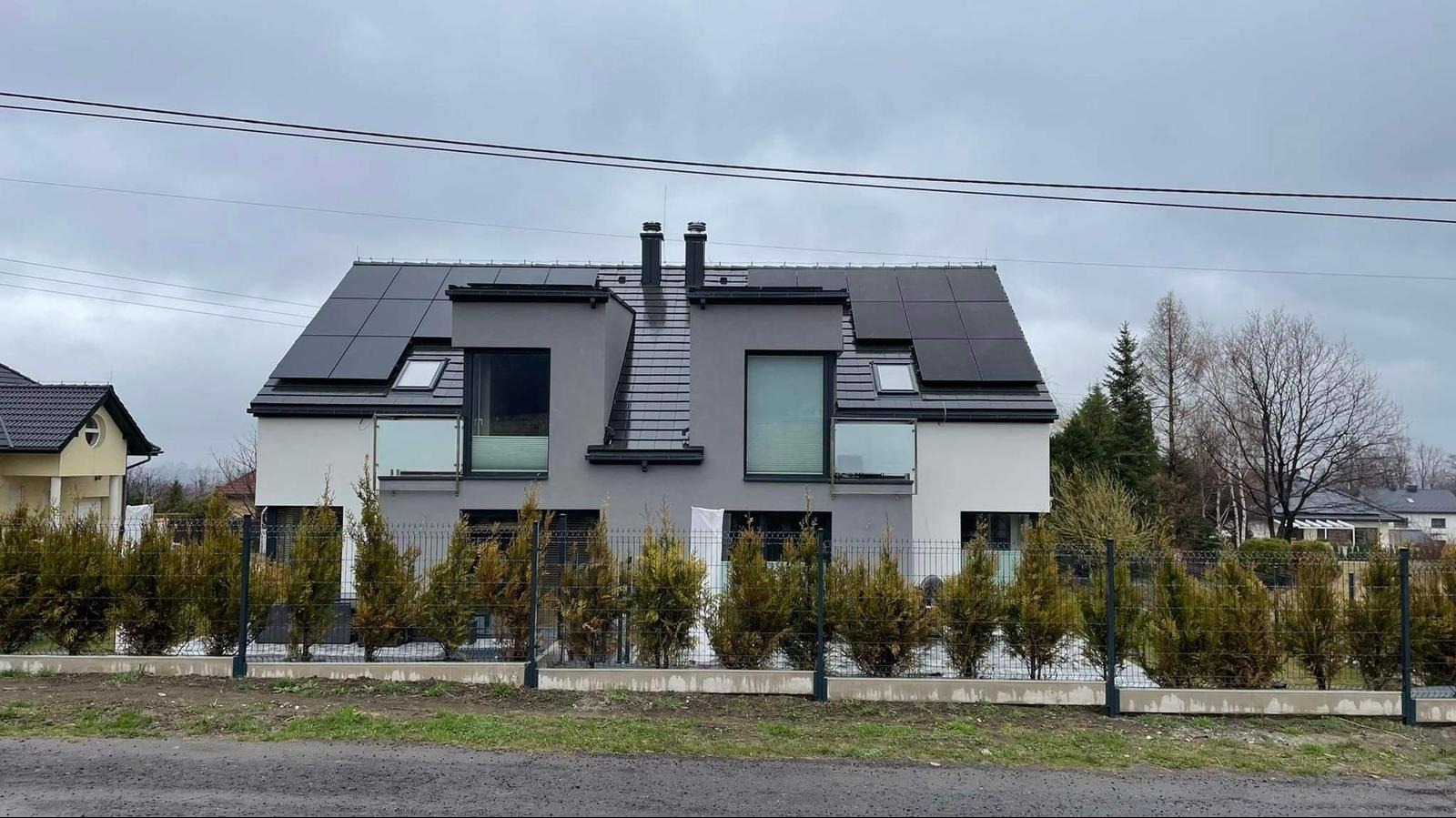 Dwukondygnacyjny dom z panelami słonecznymi na dachu, widok zza ogrodzenia i rzędu tui, pochmurne niebo w tle.
