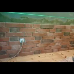 Ściana wyłożona cegłą klinkierową z widocznym gniazdkiem elektrycznym i zieloną linią lasera wyznaczającą poziom, na g&oacute;rze ściany widoczne ślady po szpachlowaniu.