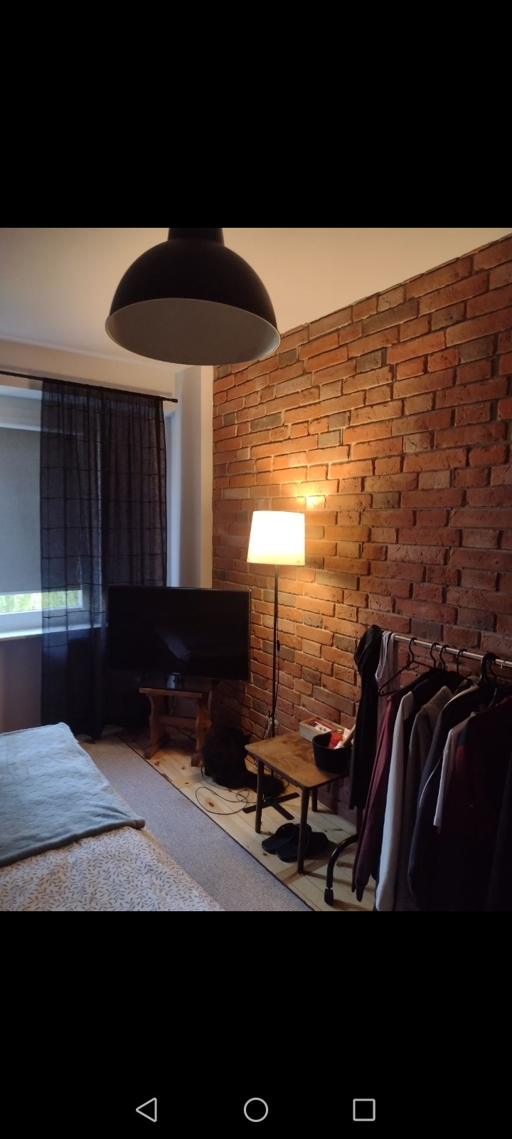 Ściana z czerwonej cegły w sypialni z elementami wystroju w stylu loft: lampa podłogowa, czarna lampa sufitowa, wieszak na ubrania.