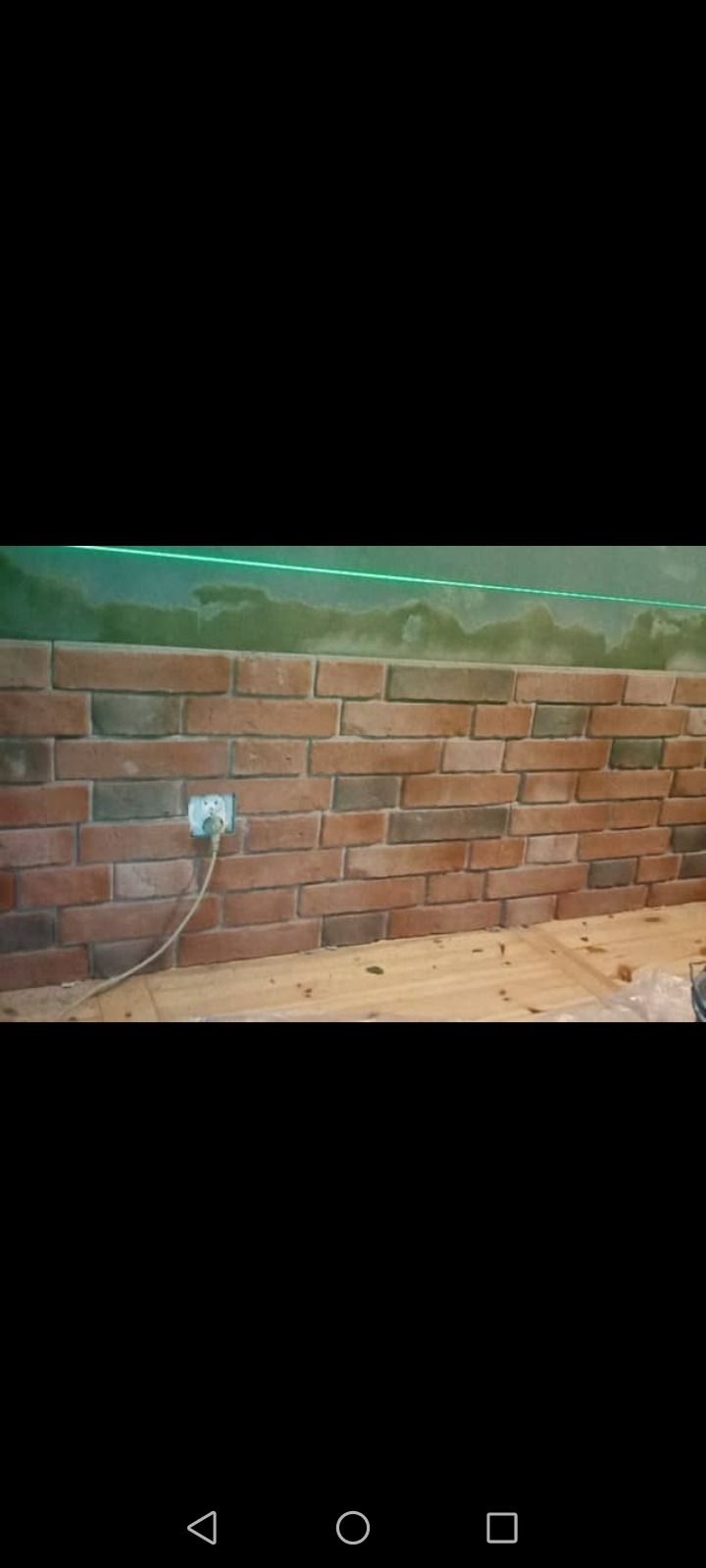 Ściana wyłożona cegłą klinkierową z widocznym gniazdkiem elektrycznym i zieloną linią lasera wyznaczającą poziom, na górze ściany widoczne ślady po szpachlowaniu.