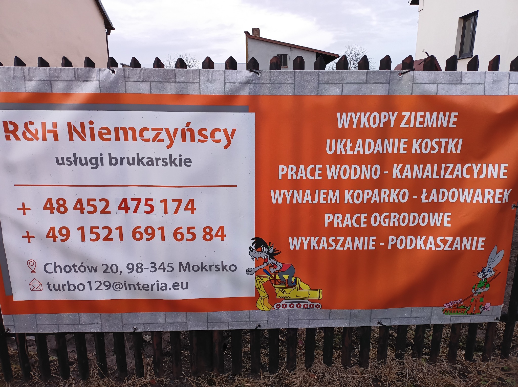 Baner reklamowy firmy R&H Niemczyńscy oferującej usługi brukarskie, wywieszony na płocie, z numerami telefonów i adresem e-mail, ilustrowany kreskówkowymi postaciami przy pracy.