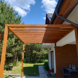 Aa bud - Drewniana pergola z belek w kolorze orzechowym, przylegająca do ściany domu z widocznym systemem rynnowym, na tle zielonego ogrodu i błękitnego nieba z chmurami.