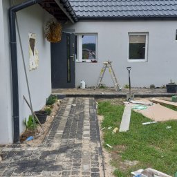 Aa bud - Ułożona częściowo kostka brukowa w odcieniach szarości prowadząca do wejścia domu z szarą elewacją, widoczne narzędzia brukarskie, drabina oparta o ścianę, teren w trakcie prac wykończeniowych.