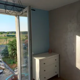 Wnętrze pokoju z otwartym oknem balkonowym, widoczna komoda z trzema szufladami, dekoracyjna ściana w odcieniach szarości i błękitu, na zewnątrz widok na ogr&oacute;d i teren budowy.