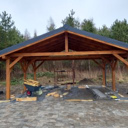 Aa bud - Drewniana konstrukcja z czarnym dachem na tle lasu, wózek z drewnem i rozłożone narzędzia na kostce brukowej, częściowo ukończona budowa.