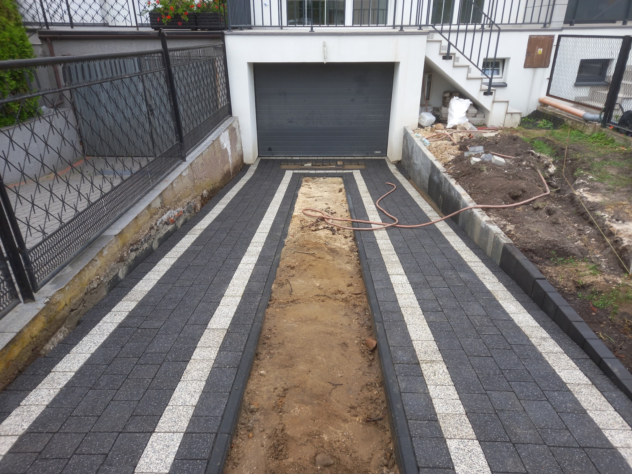 Ułożona kostka brukowa w kolorze szaro-białym prowadząca do garażu, z wykopanym pasem ziemi pośrodku, widoczne schody i barierka.