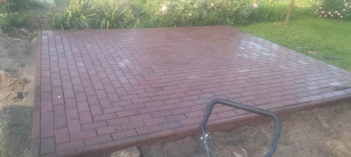 Nowo ułożona, prostokątna powierzchnia z czerwonej kostki brukowej, otoczona ziemią i trawą, z widocznym fragmentem narzędzia do układania.