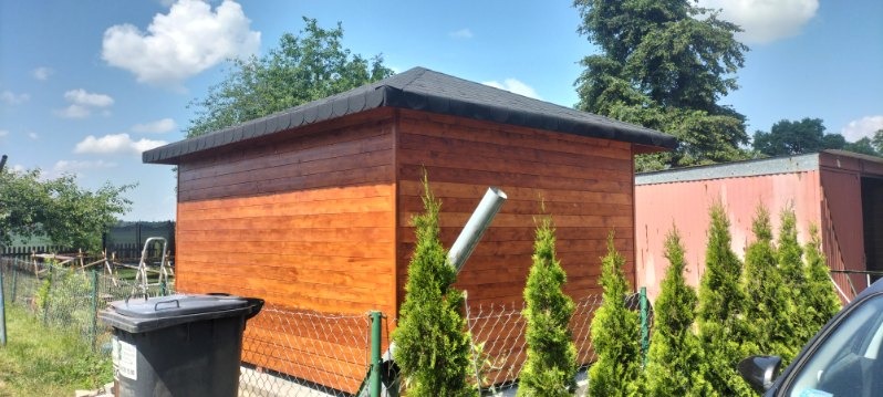 Drewniana altana z czarnym dachem, widoczny system odprowadzania wody, otoczona tujami, z koszem na śmieci i drabina w tle.