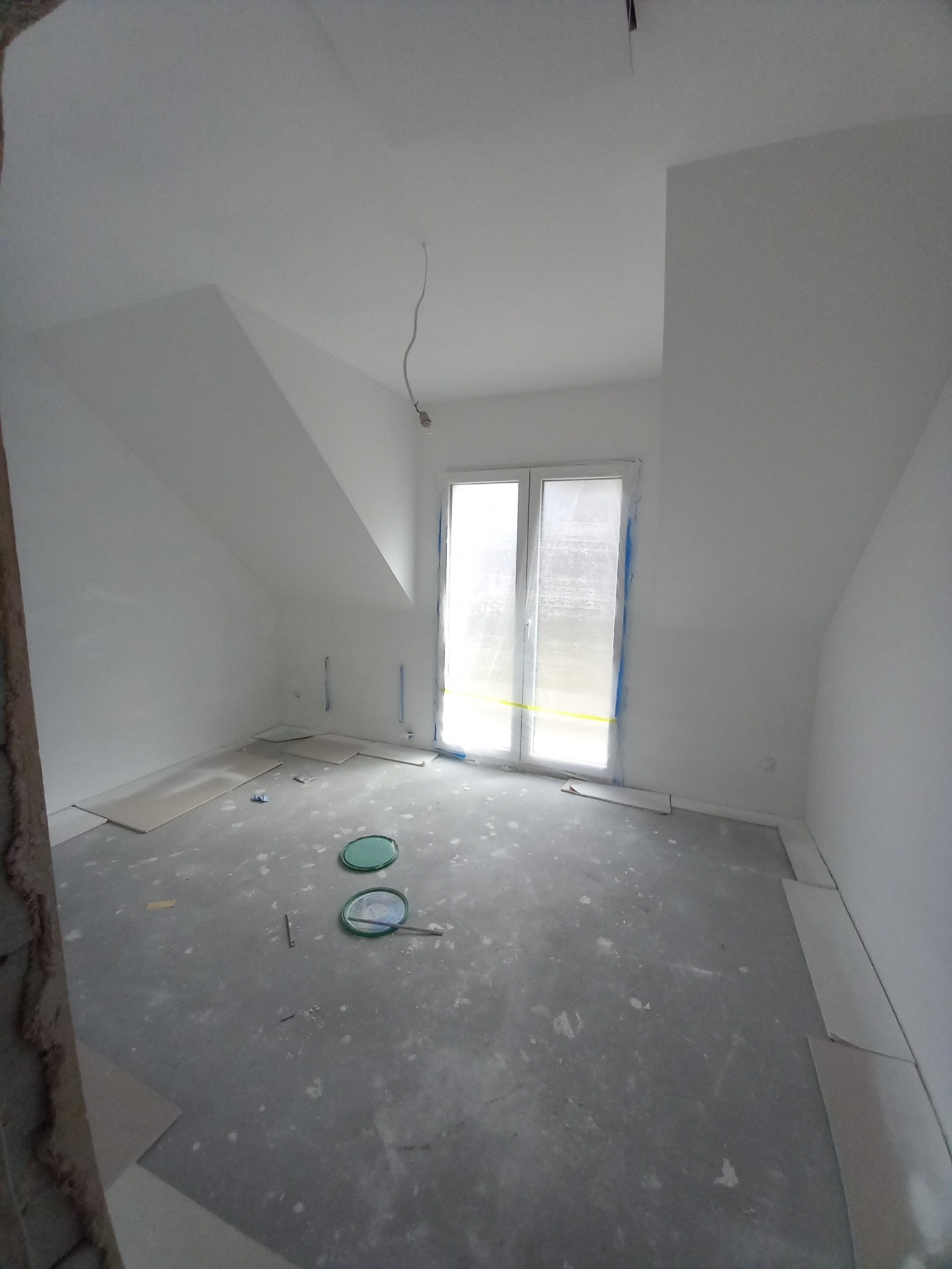 Pokój na poddaszu w trakcie remontu: świeżo pomalowane białe ściany, surowy betonowy podkład, okno zabezpieczone folią malarską, widoczne płyty gipsowe i puszki po farbie.