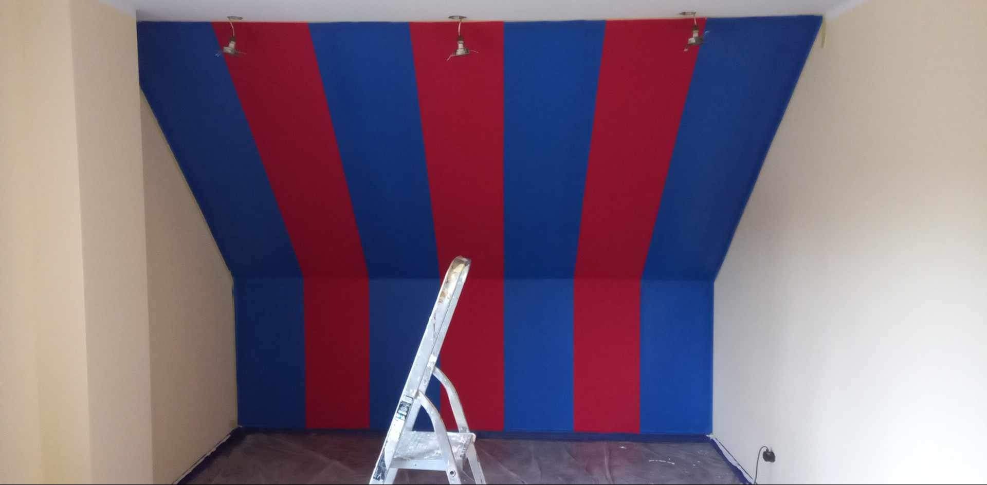 Pokój na poddaszu z pomalowanymi ścianami w pionowe pasy czerwieni i błękitu, drabina oparta o ścianę, podłoga zabezpieczona folią malarską.