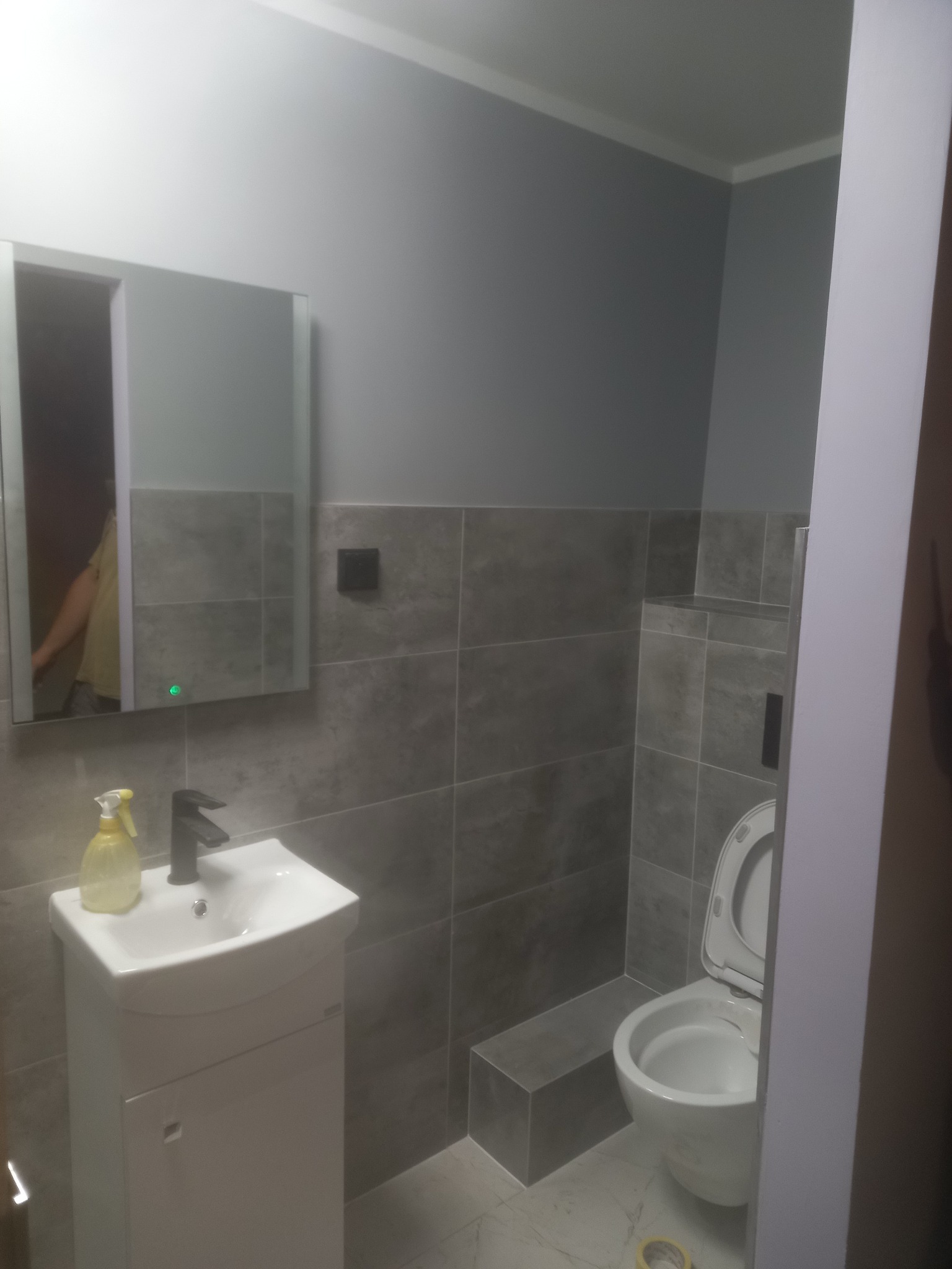 Szara łazienka z umywalką na szafce, lustrem z podświetleniem LED, toaletą i szarymi płytkami na ścianach oraz podłodze. Na umywalce stoi butelka ze spryskiwaczem.