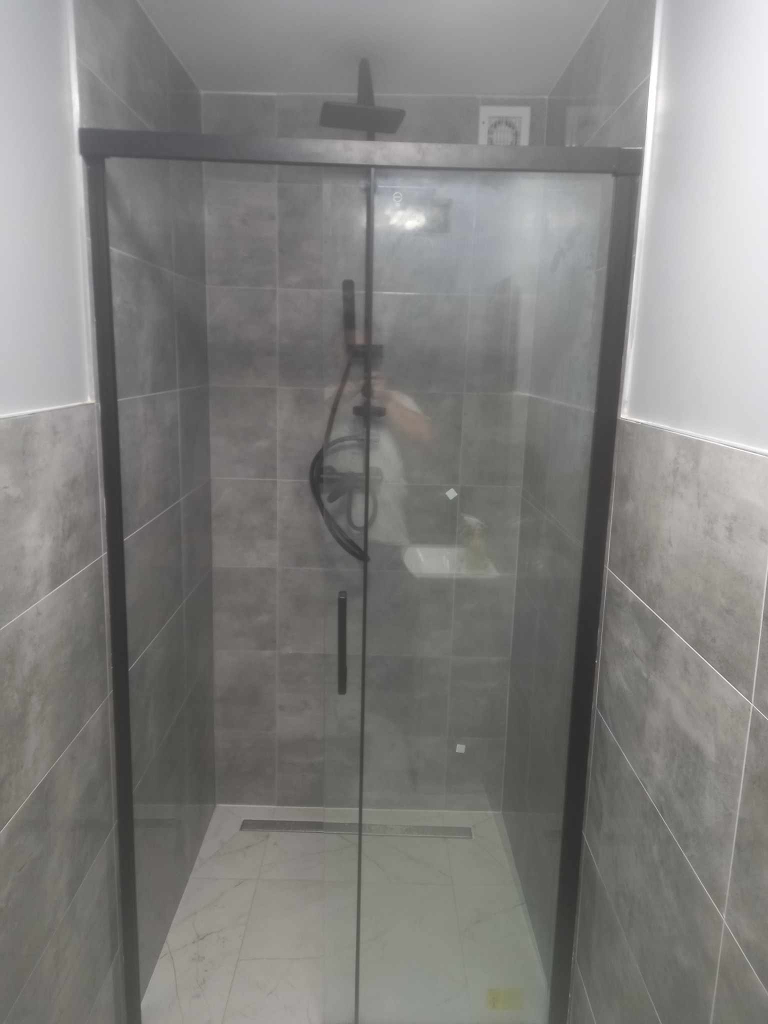 Nowoczesna kabina prysznicowa z przesuwanymi drzwiami w czarnej ramie, szare płytki na ścianach, biała marmurowa podłoga z odpływem liniowym.