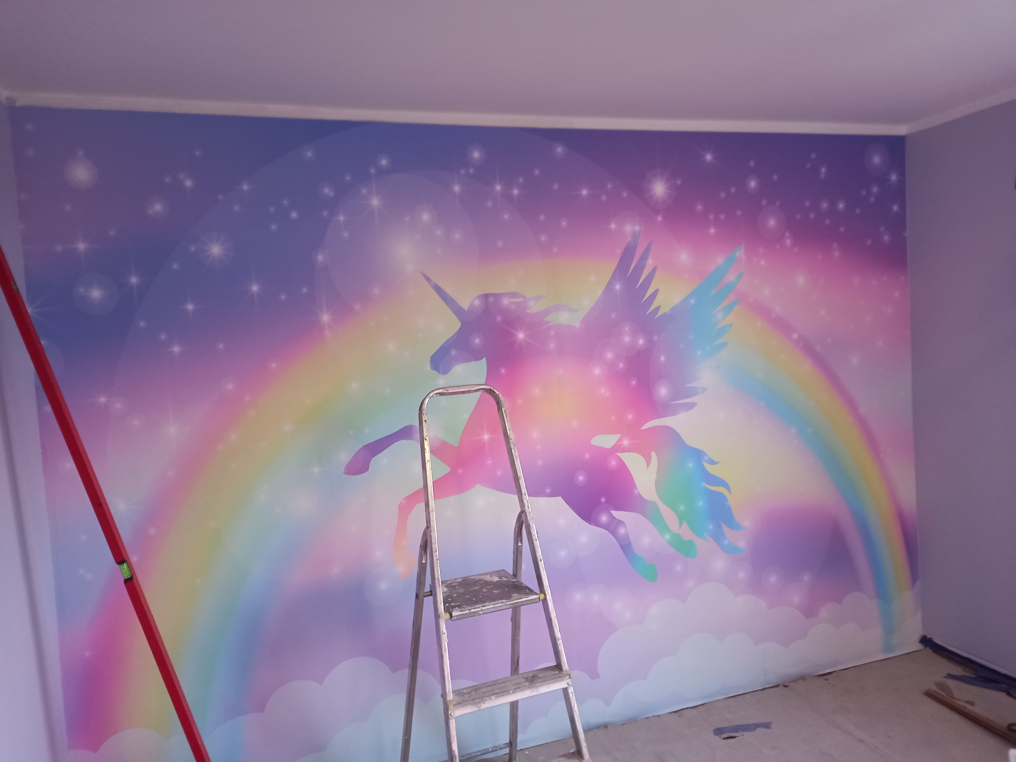 Pokój po remoncie z fototapetą przedstawiającą tęczowego jednorożca ze skrzydłami na tle gwiazd, widoczna drabina malarska i poziomica.