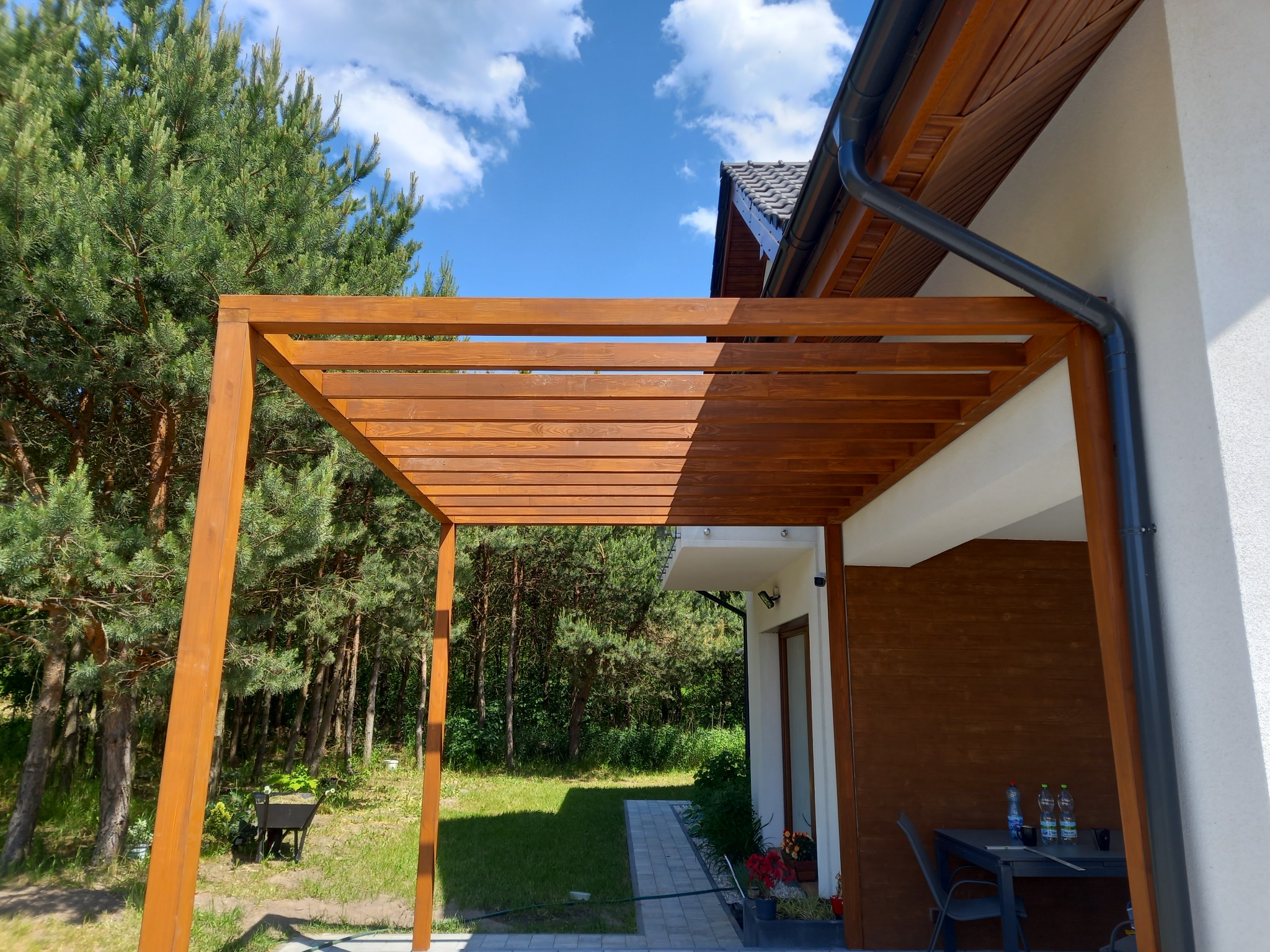 Drewniana pergola z belek w kolorze orzechowym, przylegająca do ściany domu z widocznym systemem rynnowym, na tle zielonego ogrodu i błękitnego nieba z chmurami.