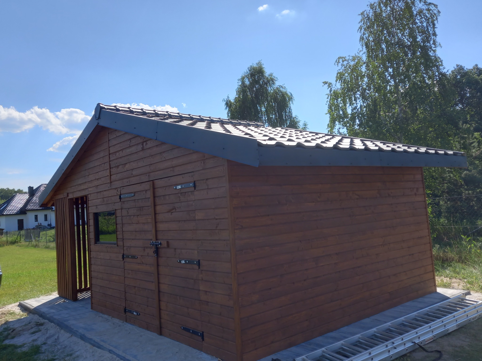 Drewniana altana z ciemnobrązowymi deskami i szarą dachówką, z drabiny opartą o bok, na tle błękitnego nieba i zieleni drzew.