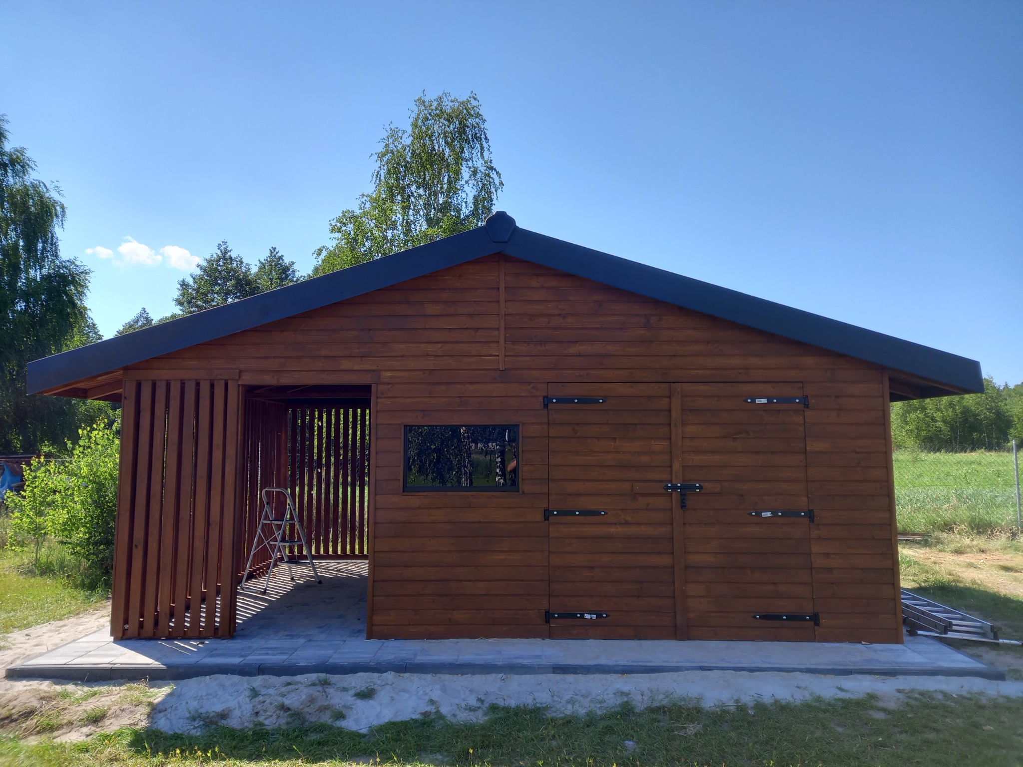 Drewniana konstrukcja z zadaszeniem, częściowo osłonięta pionowymi listwami, z drabiną wewnątrz, na tle zieleni i błękitnego nieba.