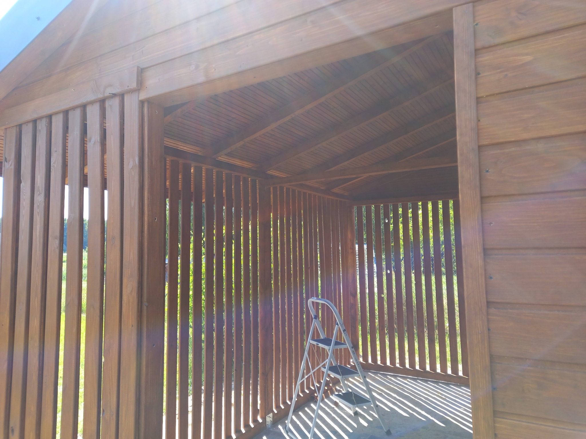 Drewniana konstrukcja altany z ażurowymi ścianami, widoczna drabina malarska wewnątrz, promienie słoneczne przebijają przez szczeliny tworząc cienie.