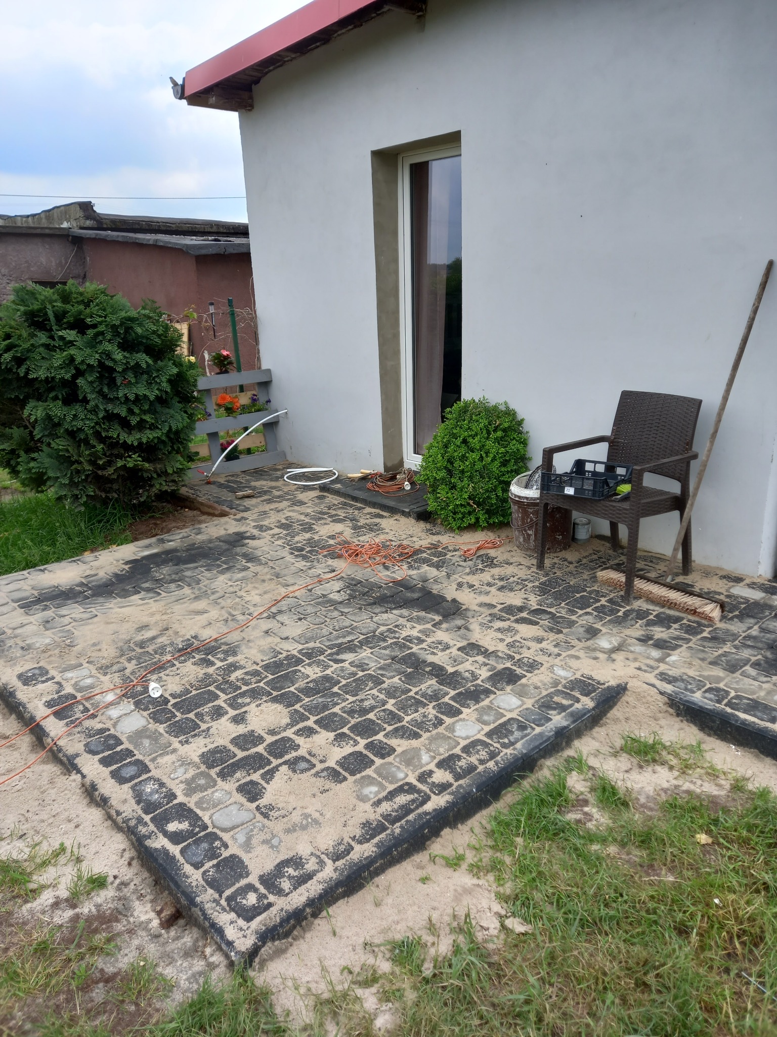 Fragment układanej nawierzchni z kostki brukowej w odcieniach szarości, z widoczną warstwą piasku i wystającym kablem zasilającym. W tle ściana budynku z oknem, krzesło z plastikową skrzynką...