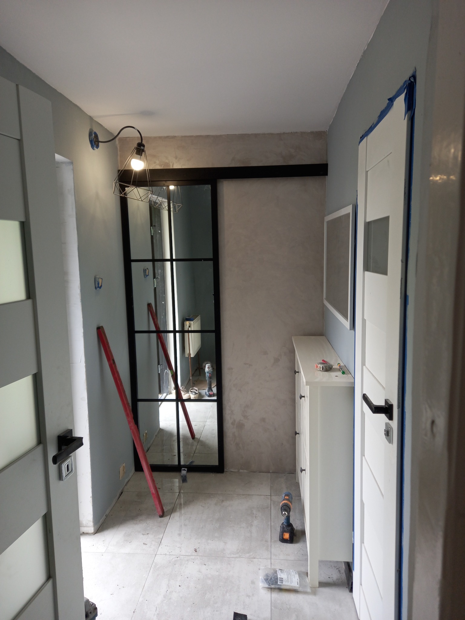 Korytarz w trakcie wykańczania: widoczne drzwi wewnętrzne z czarnymi szprosami, biała komoda z lustrem nad nią, lampa ścienna w stylu industrialnym, poziomnica oparta o ścianę, narzędzia...