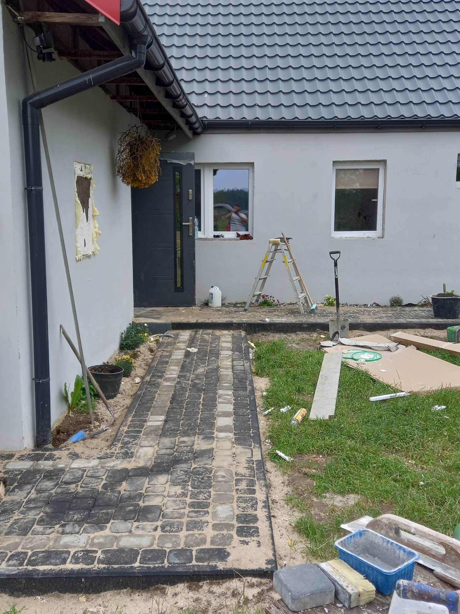 Ułożona częściowo kostka brukowa w odcieniach szarości prowadząca do wejścia domu z szarą elewacją, widoczne narzędzia brukarskie, drabina oparta o ścianę, teren w trakcie prac wykończeniowych.