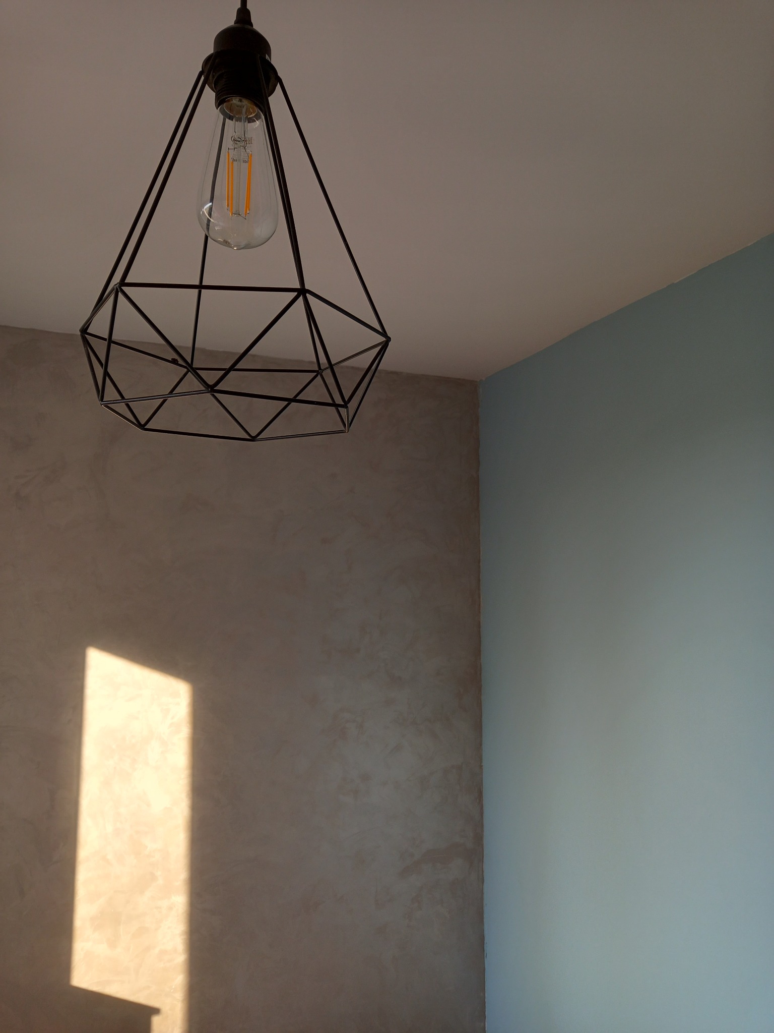 Dekoracyjna lampa wisząca w stylu industrialnym, z żarówką Edisona, oświetlająca ścianę z efektem dekoracyjnym w Bydgoszczy, obok ściana pomalowana na jasny niebieski kolor.